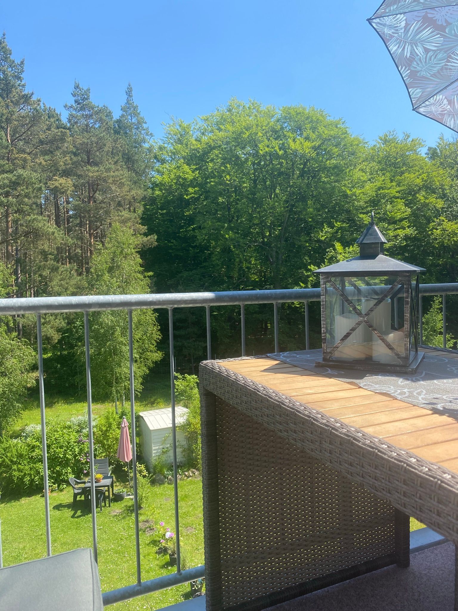 Ein Balkon mit Korbmöbeln, einer Glaskristalleuchte und Blick auf einen Garten mit Bäumen und einem Tisch mit Stühlen.