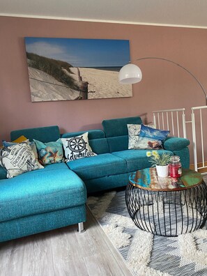 A teal sofa with multiple cushions, a round coffee table with a plant and candle, a white lamp, and a beach-themed painting on the wall.