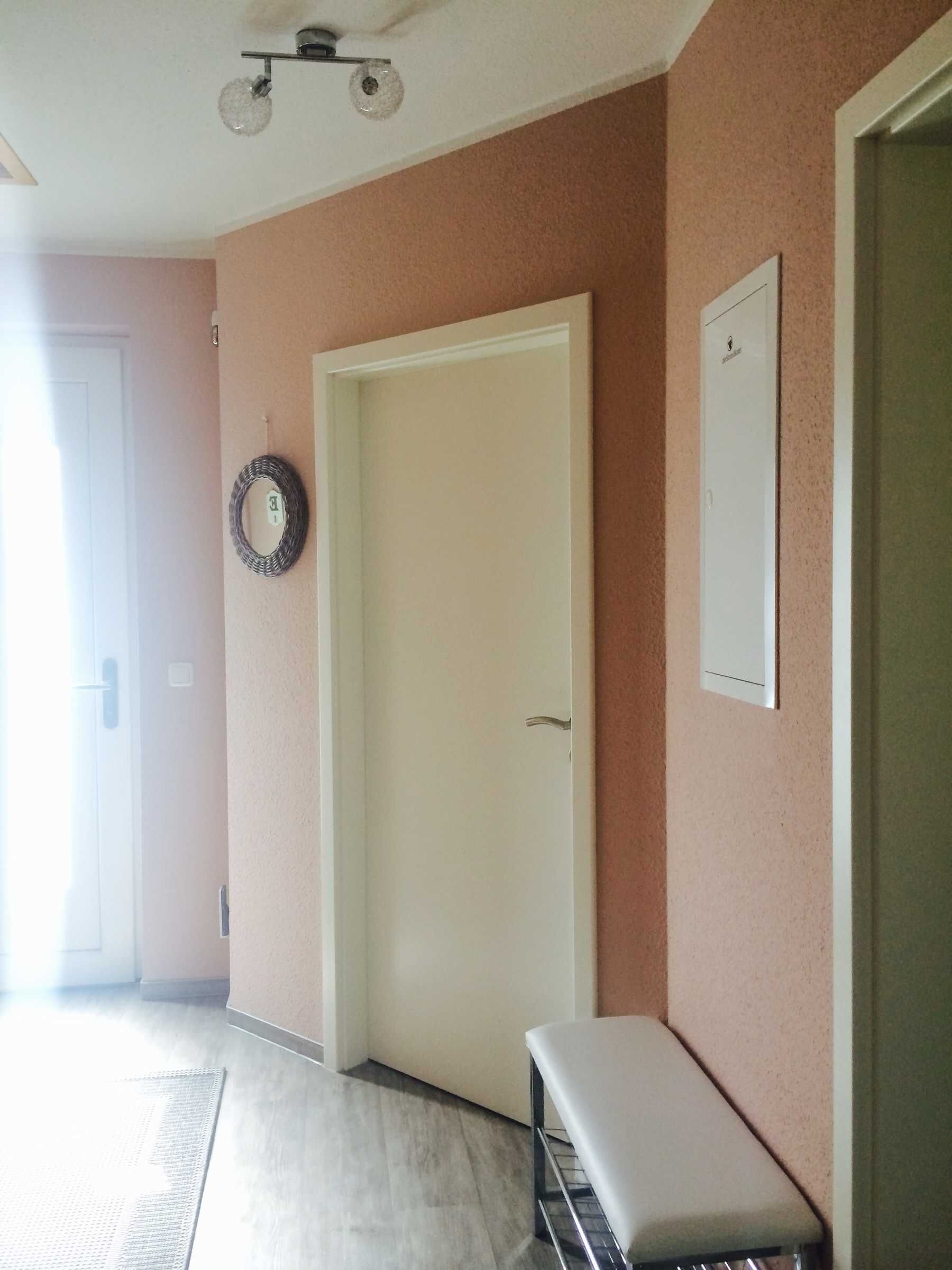 Un pasillo con una puerta blanca, un aplique en la pared y un pequeño banco.