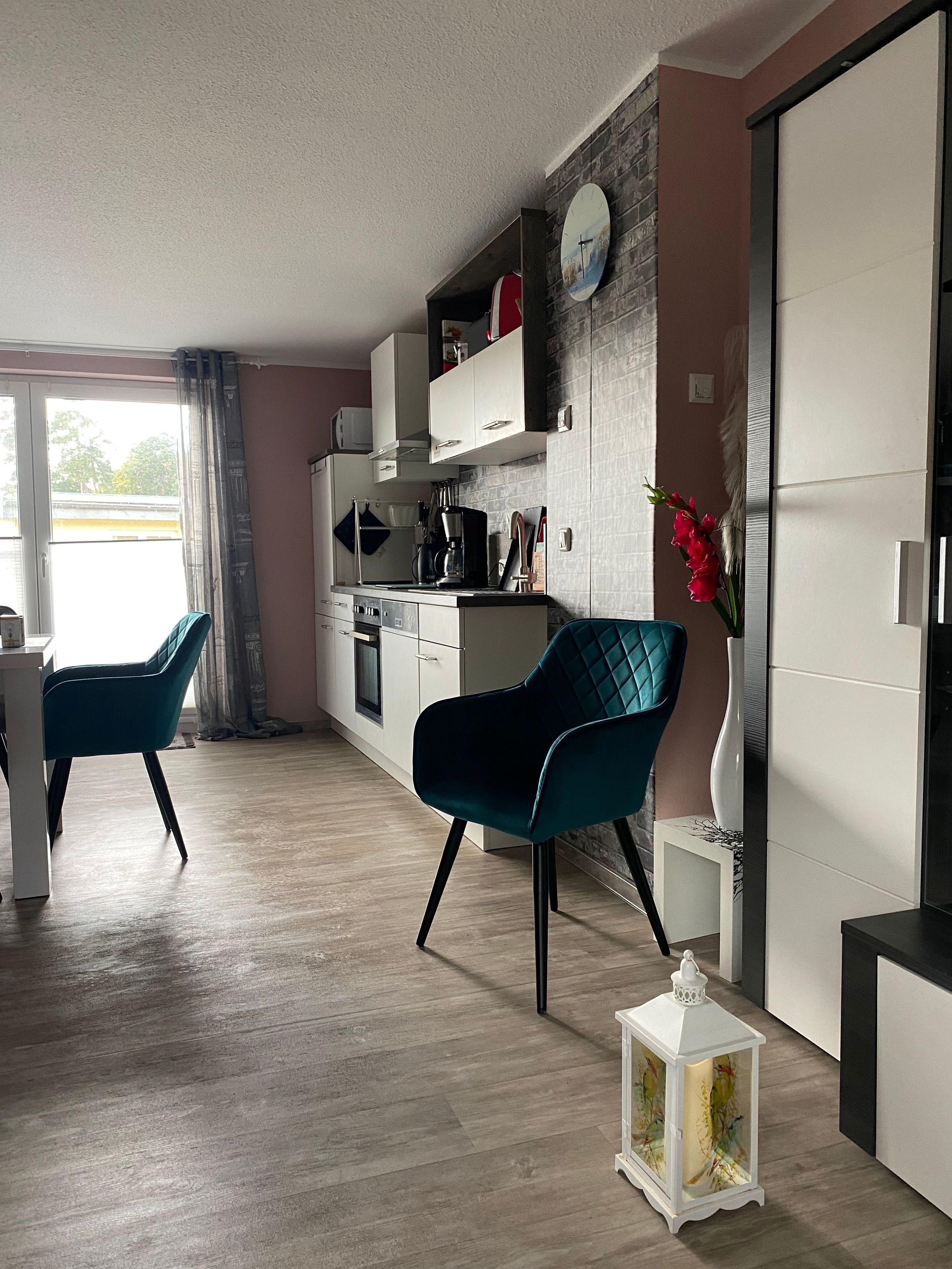 A modern kitchen with white cabinets, a dining area with teal chairs, and a white lantern on the floor.