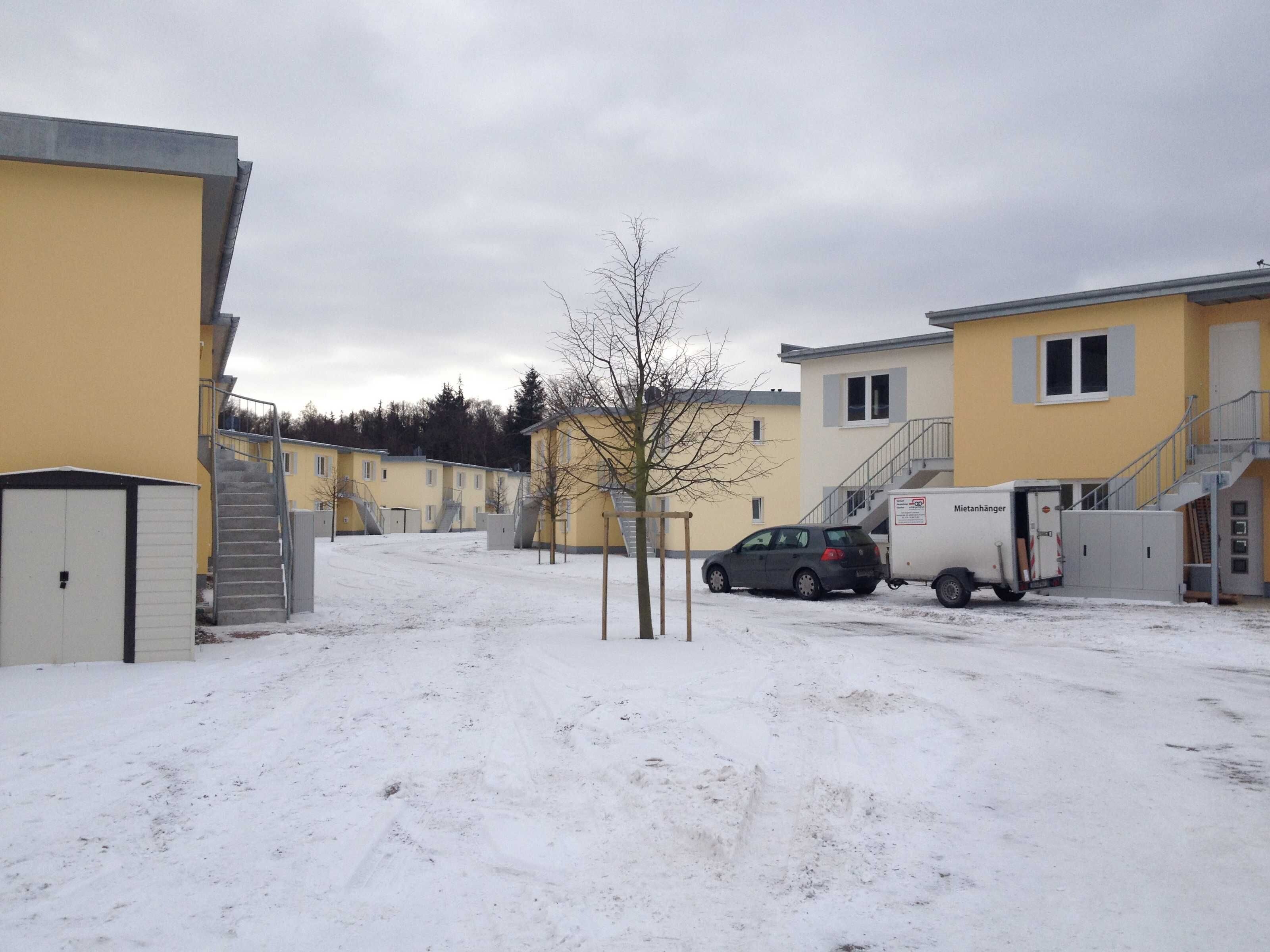 A snow-covered parking area with a car and a delivery truck, buildings with external staircases, and a bare tree.