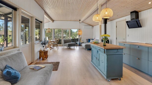 A spacious living area with a large window, a wooden ceiling, and a kitchen area with a blue cabinet and a TV mounted on the wall. - 3 bedroom amazing home in Blåvand (Blåvand)