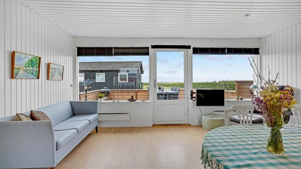 Une pièce spacieuse avec un canapé gris, une table à manger recouverte d’une nappe à carreaux et une vue sur une maison, visible à travers de grandes fenêtres.