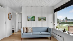Une pièce avec un canapé bleu clair, deux tableaux accrochés au mur, un miroir à cadre en bois et une fenêtre donnant sur un jardin et des maisons.