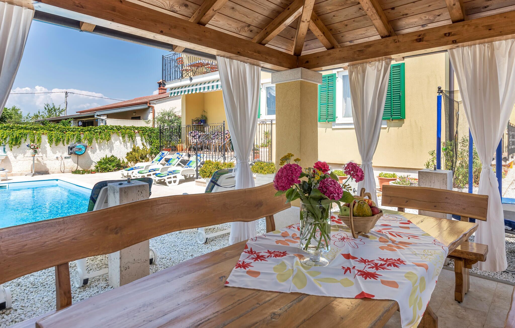 Un terrazzo in legno con un tavolo apparecchiato per mangiare, affacciato su una piscina e un giardino.