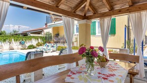 Eine Holzterrasse mit Esstisch, mit Blick auf einen Pool und einen Garten.