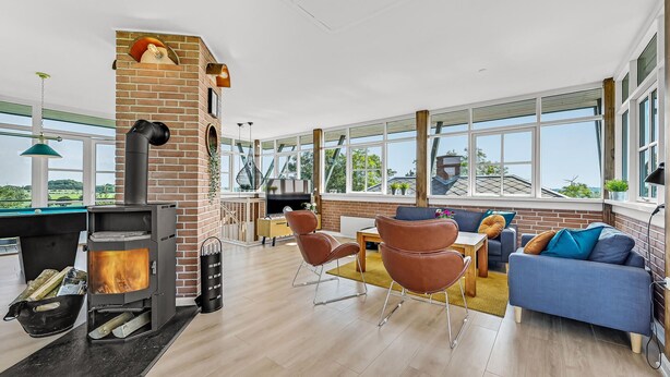 A spacious living area with a brick fireplace, modern furniture, large windows, and a view of the outdoors.