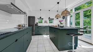 A modern kitchen with a central island, white tiled backsplash, and large windows offering a view of greenery.