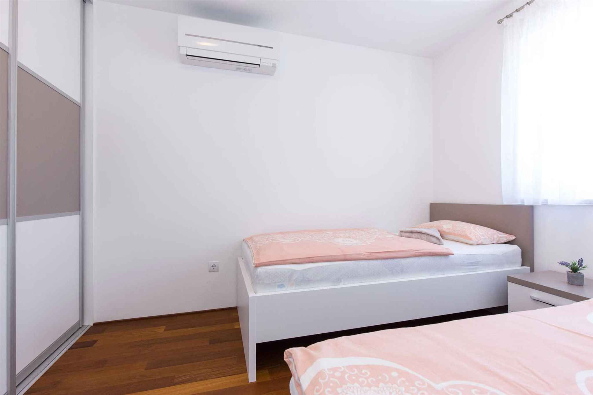 A bedroom with a bed, a bedside table, a nightstand, a window with curtains, and an air conditioning unit.