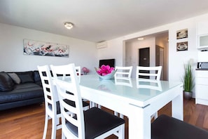 A modern living room with a glass dining table, white chairs, a dark sofa, and a painting on the wall.