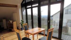 A dining area with a wooden table and four chairs, large sliding glass doors leading to a balcony with a view of a residential area. - Küpfer vacation apartment (Gräfenberg)