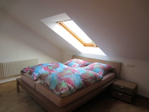 Quarto com claraboia, cama com roupa de cama colorida, mesa de cabeceira e radiador.