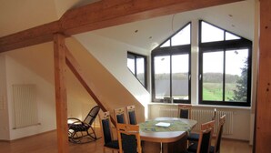 A spacious room with a high ceiling, wooden beams, a round table with chairs, and large windows offering a view of a green landscape. - Küpfer vacation apartment (Gräfenberg)
