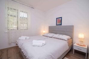 Een slaapkamer met een bed, witte gordijnen, een nachtkastje met een lamp en een lijst met een foto aan de muur.