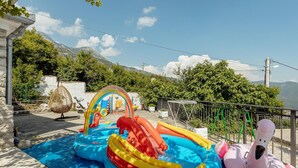 Una piscina inflable con toboganes y una pelota de playa, rodeada de árboles y un paisaje montañoso de fondo.