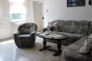 Ein Wohnzimmer mit einer gemusterten Couch, einem kleinen runden Tisch mit Vase und Fernbedienung sowie einer Stehlampe.