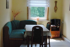 Ein Zimmer mit einem grünen Sofa, einem Tisch mit weißem Tischtuch, einem Holzstuhl, einem Fenster mit Blick auf Bäume und einer Holzkommode mit einem Fernseher.