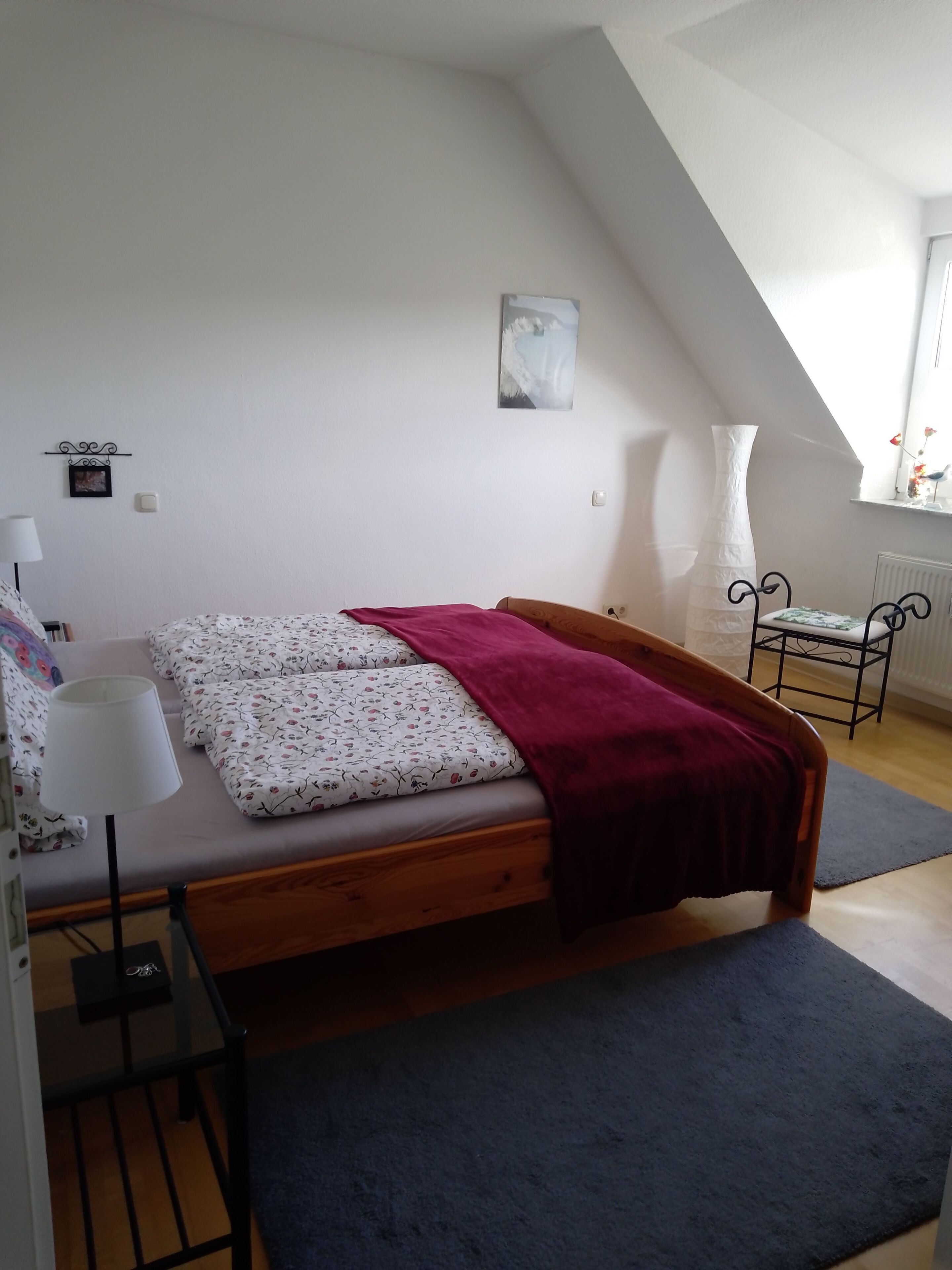 A bedroom with a wooden bed, a red blanket, a white vase, and a painting on the wall.
