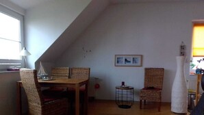 Une pièce chaleureuse de style loft, avec un plafond en pente, une table à manger entourée de chaises en osier, une petite table d’appoint et un tableau encadré accroché au mur.