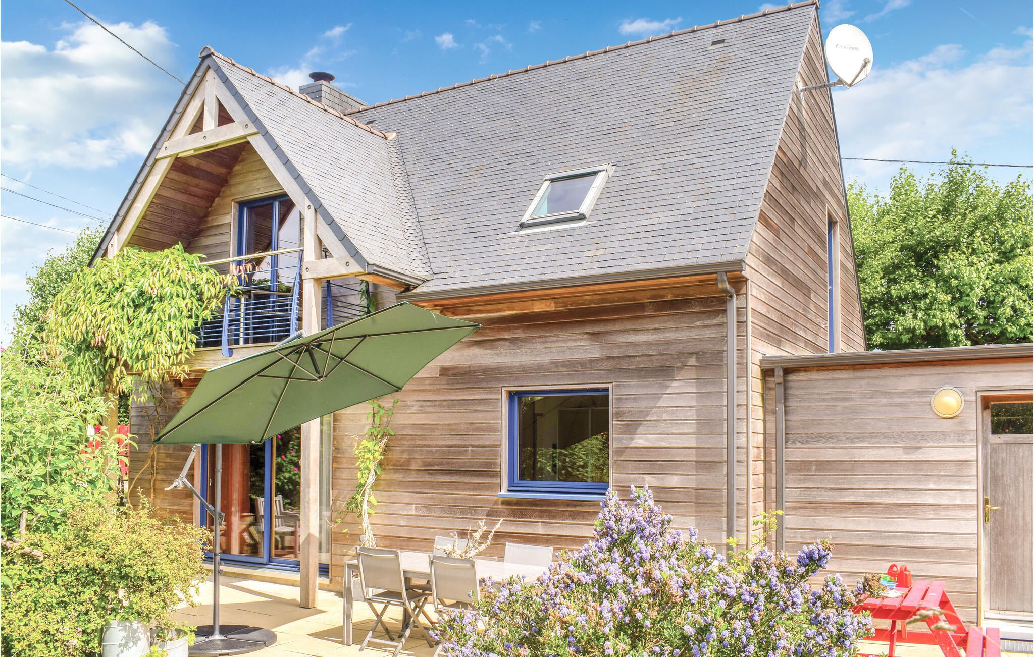 Une maison en bois à deux étages, avec un toit en pente, une antenne parabolique et une terrasse aménagée avec du mobilier d’extérieur.