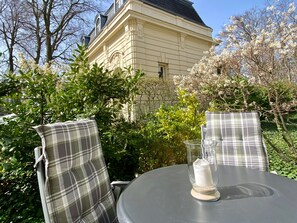 An outdoor table with two plaid chairs and a glass candle holder, set against a backdrop of a building and blooming trees.