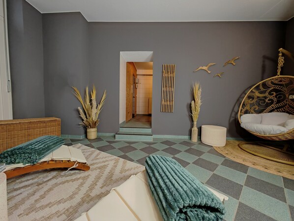 A room with a checkered floor, a bed with a white and teal blanket, a wicker chair with a white cushion, and a hanging chair with a patterned design.