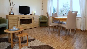 Una sala de estar con un mueble de madera, un televisor, un sofá verde, una mesa de comedor con sillas y una ventana con cortinas.