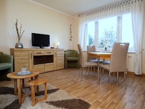 Ein Wohnzimmer mit Holzschrank, Fernseher, einem grünen Sofa, einem Esstisch mit Stühlen und einem fenster mit Vorhängen.