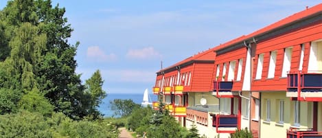 A row of red and yellow buildings with balconies, a paved walkway, and a sailboat in the background.