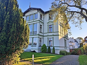 A multi-story building with a classical architectural style, featuring a prominent balcony and a well-maintained garden area.