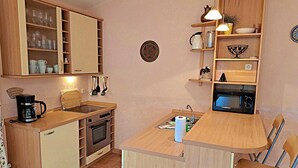 Cocina compacta con armarios de madera, horno integrado, cafetera y un pequeño comedor con una silla.