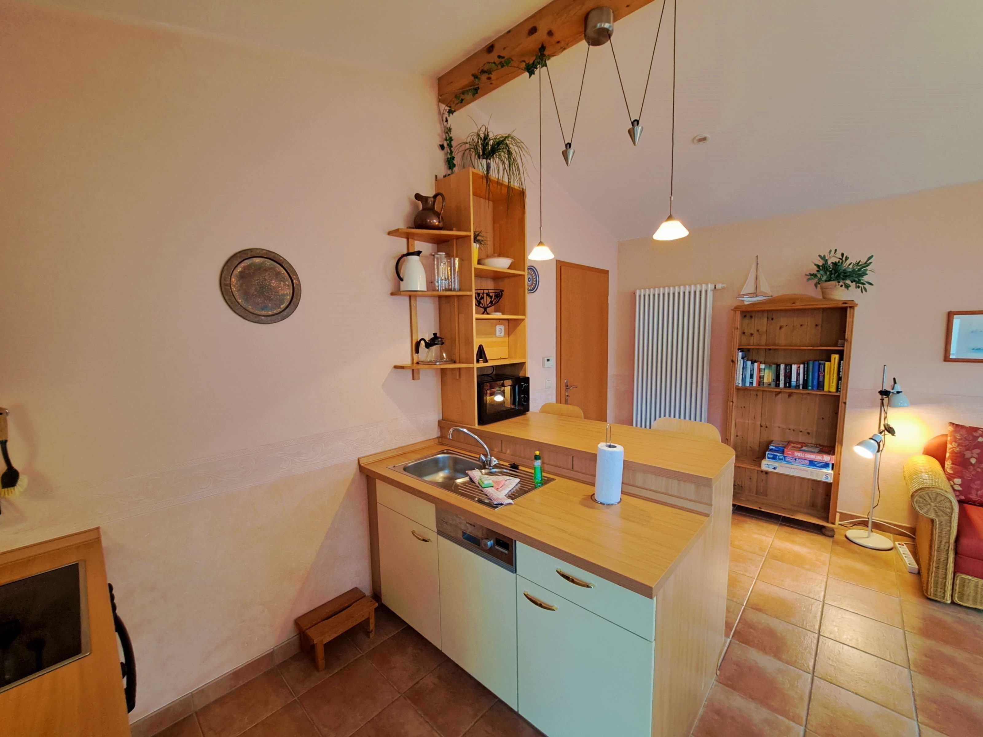A compact kitchen with a sink, wooden cabinets, and a small dining table. There is a bookshelf filled with books and a potted plant.