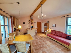 A cozy living room with a wooden dining table, wicker chairs, a red sofa, and a kitchen area with wooden cabinets and a hanging light fixture.