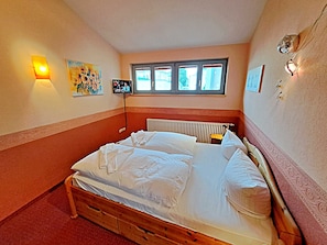 Ein Schlafzimmer mit einem hölzernen Bett, einem Heizkörper, einem an der Wand montierten Fernseher und einem Fenster mit Jalousien.
