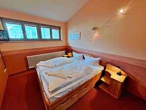 Une chambre à coucher avec un lit en bois, une petite table de chevet, un radiateur et une fenêtre donnant sur des immeubles.