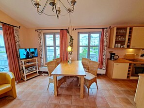 A cozy living room with a wooden dining table, wicker chairs, a television, and a kitchen area with cabinets and a coffee maker.