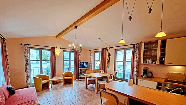 Una sala de estar acogedora con sofá, sillas, una mesa de comedor y televisión. Hay ventanas amplias con cortinas, una zona de cocina con armarios y una lámpara colgante.