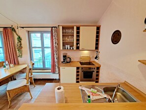 A compact kitchen with a dining area, featuring a wooden table, wicker chairs, a built-in oven, and a window with curtains.