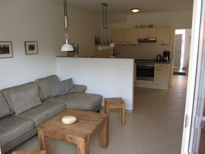 Sala de estar com um sofá cinza, uma mesa de centro de madeira e um banquinho de madeira pequeno. Há uma cozinha ao fundo, com armários e um fogão.