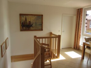 Una escalera con barandillas de madera que conduce a un rellano, un cuadro de un barco velero en la pared, una puerta y una ventana con cortinas.