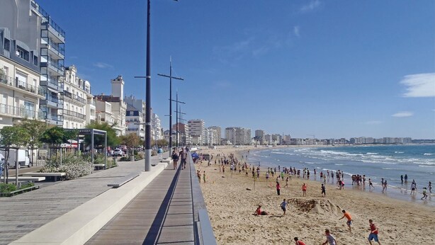 Una playa con un paseo marítimo de madera, gente jugando y edificios al fondo.