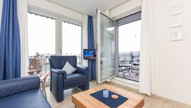 Ett modernt vardagsrum med en soffa, fåtölj och soffbord, stora fönster med gardiner och en tv som står på ett tv-bord.