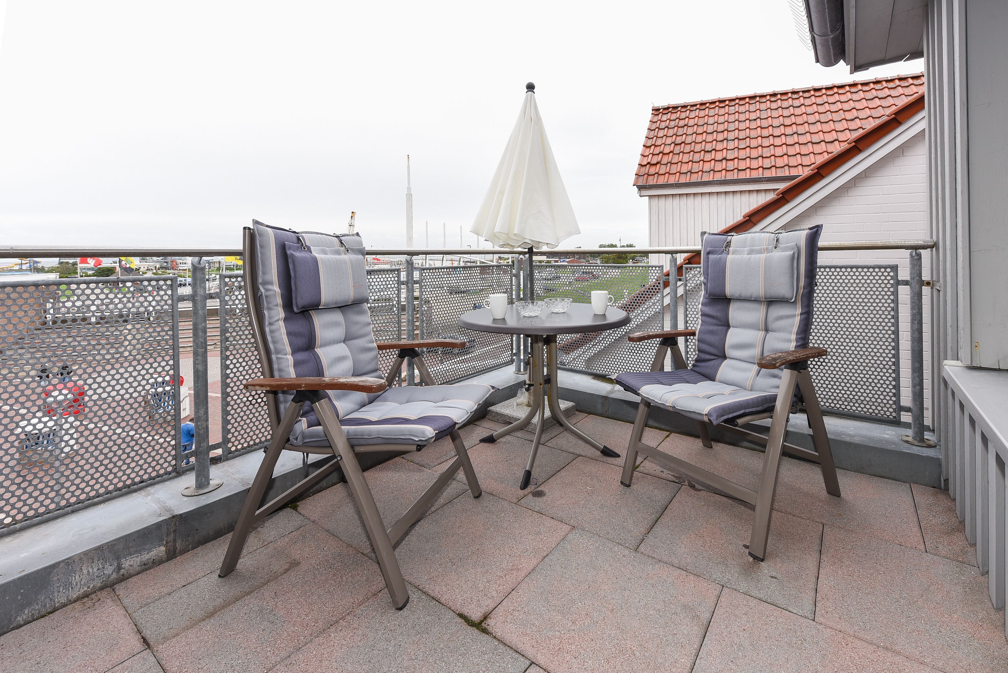 Ein Balkon mit zwei Stühlen, einem kleinen Tisch, einem weißen Sonnenschirm und Blick auf ein Gebäude mit rotem Dach.