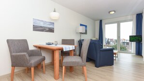 Ett modernt vardagsrum med ett matbord, två grå fåtöljer, en blå soffa, en tv och ett stort fönster med gardiner.
