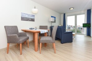 Un salon moderne comprenant une table à manger, deux fauteuils gris, un canapé bleu, un téléviseur et une grande fenêtre avec des rideaux.