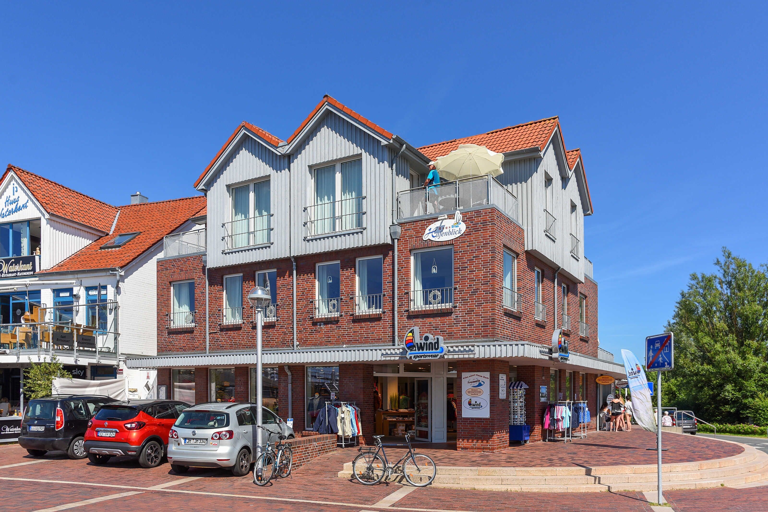 Un immeuble à plusieurs étages avec une façade en briques, un balcon agrémenté d’un parasol rouge et une devanture de magasin sur laquelle on peut lire : « Wind Apartments ».