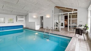 An indoor swimming pool with clear blue water, a blue poolside chair, and a dining area with a table and chairs. - Amazing home in Blåvand with sauna (Blåvand)