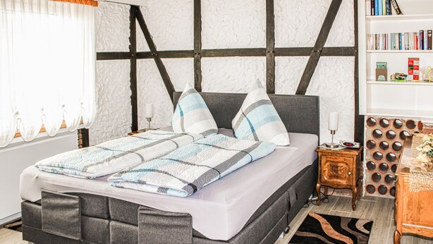 A modern bedroom with a grey upholstered bed, patterned bedding, a wooden nightstand, and a bookshelf with books and decorative items. - Beautiful apartment in Reifferscheid (Reifferscheid)