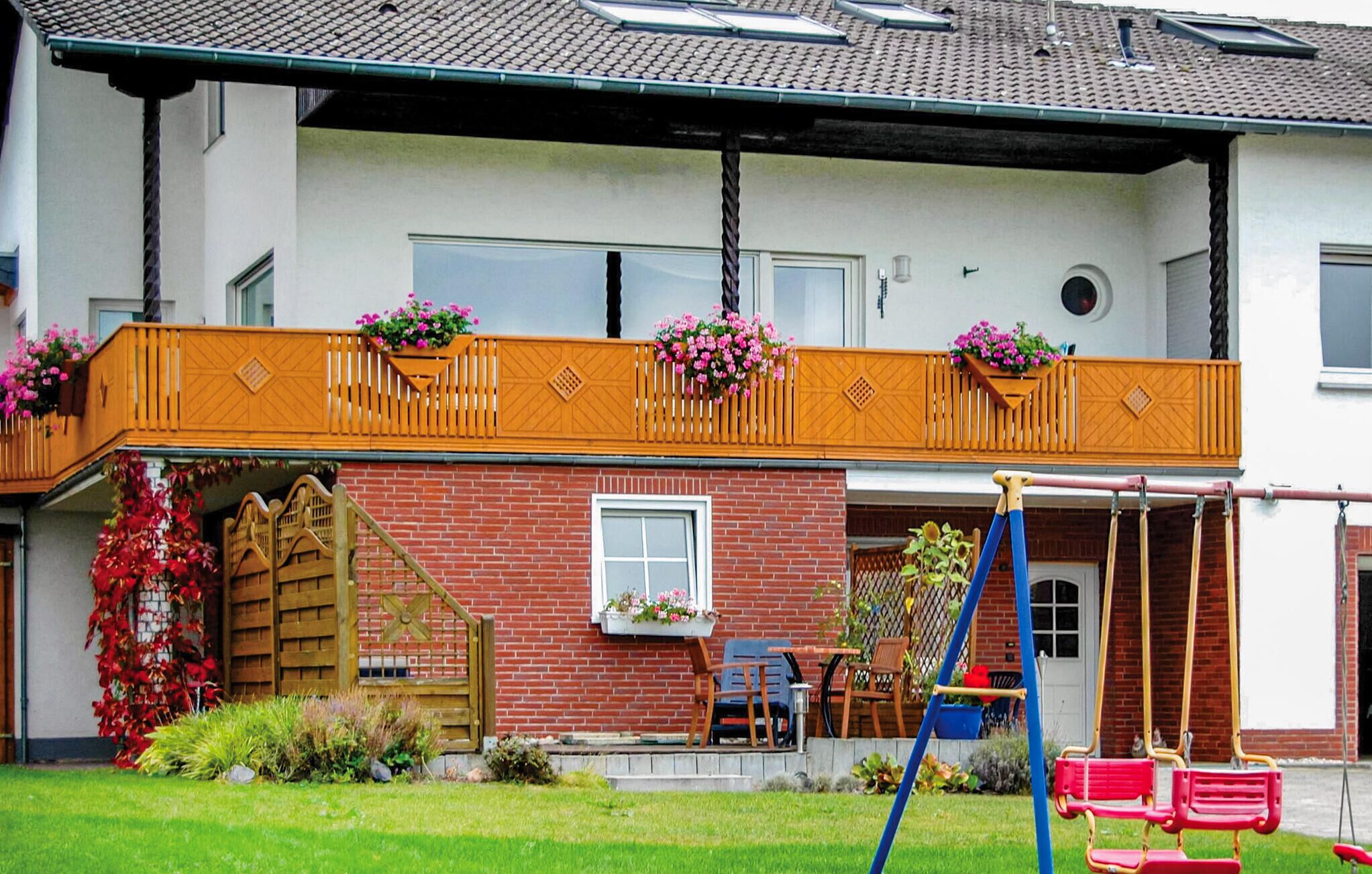 A two-story house with a red brick facade, a wooden balcony with flower boxes, a swing set, and a wooden gate.