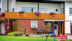 A two-story house with a red brick facade, a wooden balcony with flower boxes, a swing set, and a wooden gate. - Beautiful apartment in Reifferscheid (Reifferscheid)